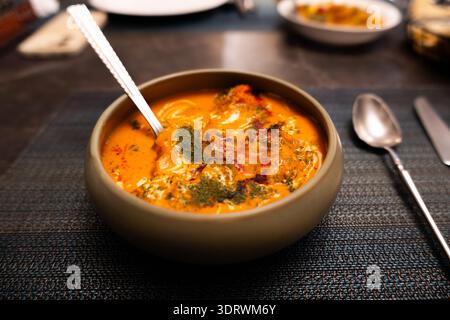 Indische Curry-Suppe in Tonschale mit Löffel auf dunkler Tischdecke, garniert mit Kräutern und Gewürzen Stockfoto
