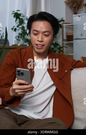 In einer stilvollen Wohnung lächelt ein junger asiatischer Mann hell, während er auf seinem Smartphone blättert. Stockfoto