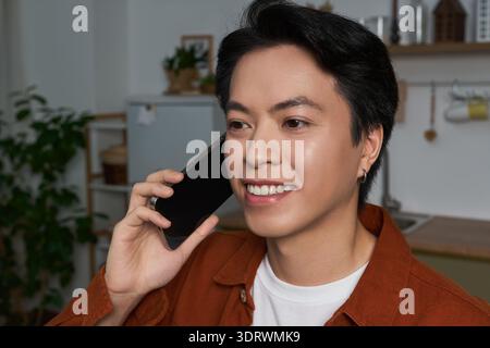 Ein asiatischer Mann lächelt hell, während er in einem gemütlichen Wohnraum mit seinem Telefon spricht. Stockfoto