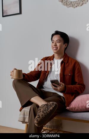 Lässig gekleidet sitzt ein junger asiatischer Mann bequem in seiner gemütlichen Wohnung und schlürft Tee. Stockfoto