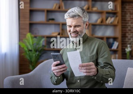 Lächelnder reifer Mann, der einen Papierbeleg mit einem Mobiltelefon scannt, um persönliche Finanzen zu verwalten, Ausgaben zu verfolgen oder eine digitale Banking-Anwendung zu Hause zu nutzen Stockfoto
