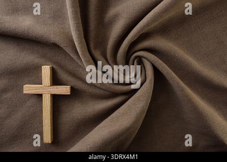 Ein hölzernes Kreuz, das auf einem braunen Stoff aufliegt und einen warmen, minimalistischen christlichen Hintergrund mit eleganter Textur und viel Kopierraum bildet. Stockfoto