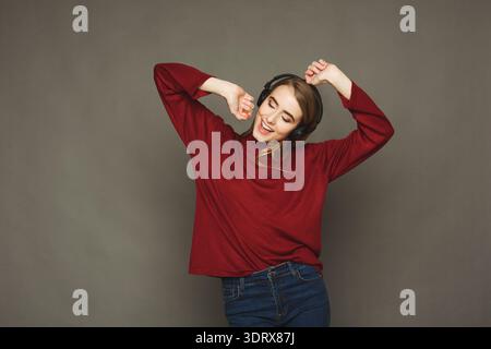 Junge Frau tanzt glücklich, während sie Kopfhörer in einer einfachen Innenumgebung trägt Stockfoto