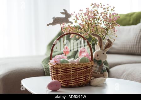 Osterkorb mit Ester-Eiern, Plüschhase und Frühlingsdekor auf weißem Tisch im Cozy Wohnzimmer Stockfoto