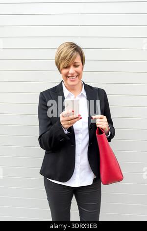 Erwachsene weiße Geschäftsfrau in schwarzem Blazer lächelt beim Blick auf das Smartphone, steht vor neutraler Wand, Kommunikationskonzept. Stockfoto