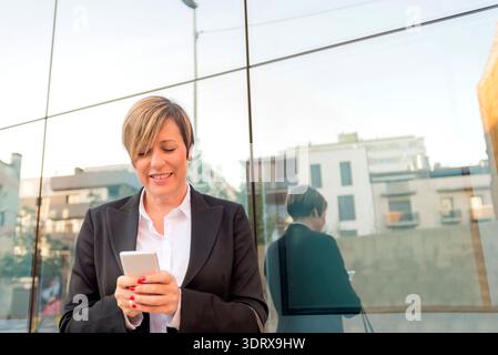 Erwachsene weiße Geschäftsfrau mit schwarzem Blazer steht draußen in der Nähe der Glaswand mit Smartphone und lächelnd. Urbane Umgebung. Stockfoto