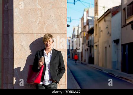 Erwachsene weiße Geschäftsfrau in Blazer mit Kaffee und roter Tasche, stehend in der städtischen Gasse. Professioneller Lebensstil und Pendelkonzept. Stockfoto