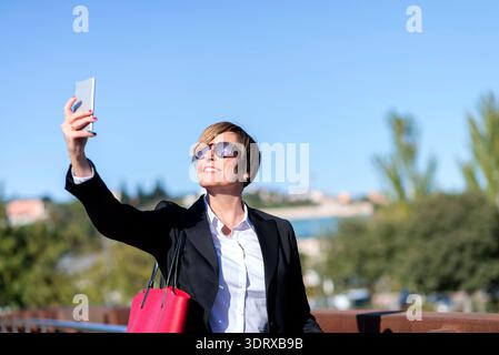 Erwachsene weiße Geschäftsfrau in schwarzem Anzug und Sonnenbrille macht Selfie mit Smartphone draußen auf Brücke, urbanem Hintergrund und sonnigem Himmel. Stockfoto