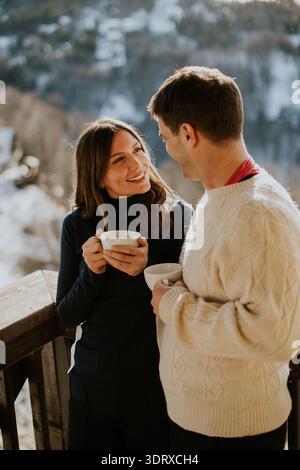 Zwei Personen teilen sich ein Lächeln bei Dampfbechern, während sie sich auf einem gemütlichen Balkon entspannen, der im Winter von schneebedeckten Bergen umgeben ist. Stockfoto
