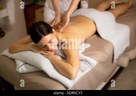 Eine Frau liegt auf einem Massagetisch in einem Wellnesscenter. Eine Therapeutin übt leichten Druck auf ihren Rücken aus. Das Zimmer hat eine ruhige Atmosphäre mit sanftem Licht Stockfoto