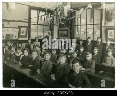 Schulkinder der 1900er 1930er Jahre, die mit dem Klassenlehrer im Klassenzimmer sitzen. Schüler der 1900er 1930er Jahre - Mädchen in der Junior-Schule, Jungs sitzen, Schreibtischreihen Großbritannien Stockfoto