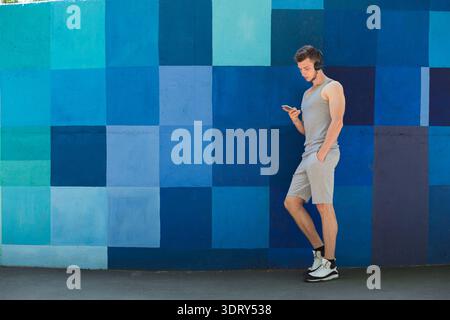 Junger Mann in grauem Outfit hört Musik, während er neben der farbenfrohen Mauer steht Stockfoto