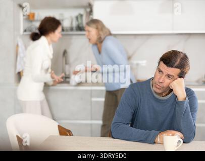 Die alte Frau streitet mit der Frau, während ein trauriger Mann mit ihnen in der Küche sitzt Stockfoto