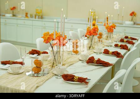 Ein Esstisch ist für ein Treffen eingerichtet. Es verfügt über orangefarbene Blumen, Kerzen und dekorative Kürbisse. Weiße Stühle umgeben den Tisch, der sich befindet Stockfoto