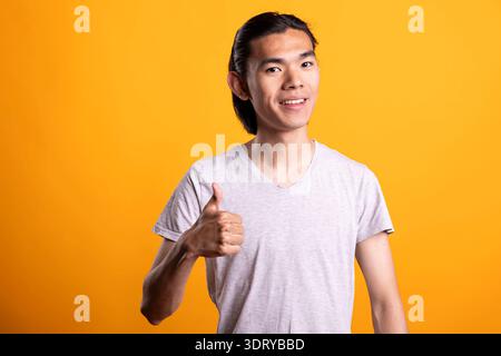 Asiatischer Mann posiert vor isoliertem Hintergrund, trägt lässiges Outfit und zeigt selbstbewusste Daumen nach oben. Das hübsche Modell lächelt hell und drückt Zufriedenheit und erfolgreiche Zustimmung bei Porträtaufnahmen aus. Stockfoto
