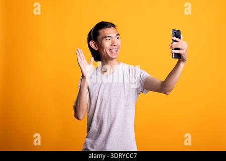 Aufgeregtes männliches Model, das fröhlich dem Handy zuwinkt und sich im Studio aufnimmt. Fröhlicher gutaussehender Blogger spricht mit Online-Zuschauern oder Familie durch herzliche und freundliche Begrüßung auf der Telefonkamera. Stockfoto