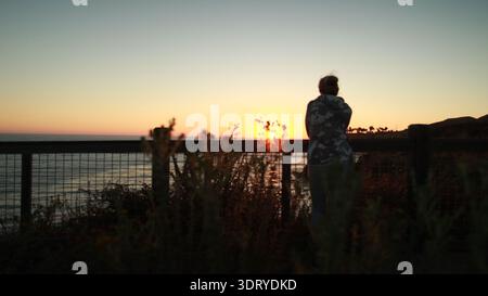 Eine weibliche Silhouette in einem Camouflage-Pullover lehnt sich an einen Zaun und beobachtet den Sonnenuntergang über dem Pazifik. Die goldene Stundenanzeige leuchtet auf Stockfoto