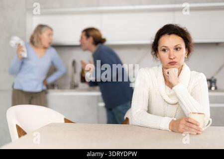 Traurig denkende Frau sitzt in der Küche und hört dem Streit zwischen Mann und älterer Frau zu Stockfoto