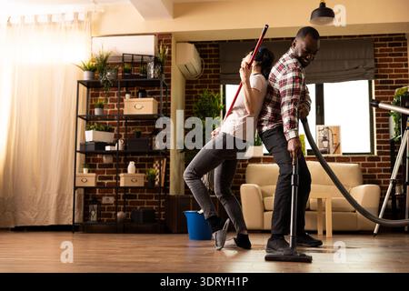 Die kaukasische Frau lehnt sich auf den Ehemann und gibt vor, Mopp-Stick als Gitarre zu spielen, während sie mit Kopfhörern singt. Afroamerikanischer Mann saugt Boden, während ein vielseitiges Paar Spaß beim Housecleaning macht. Stockfoto