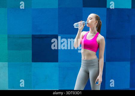 Eine junge Frau macht eine Pause und trinkt während des Workouts in der Nähe einer Blauen Mauer in urbaner Umgebung Wasser Stockfoto