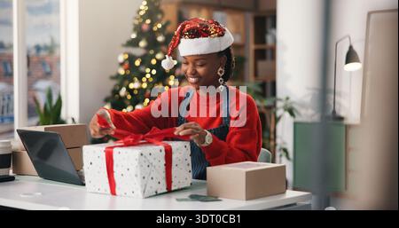 Weihnachten, Geschenk und Verpackung mit schwarzer Frau im Büro für Feiertage, Startup oder Laptop. Weihnachtszeit, Geschenk und Dropshipping mit Stockfoto