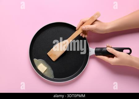 Weibliche Hände halten die Pfanne mit Butter und Spachtel auf rosafarbenem Hintergrund Stockfoto