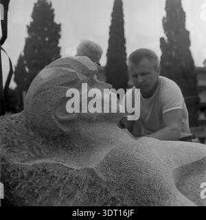 Ein historisches Archivfoto aus dem Jahr 1974, das den detaillierten Prozess der Schnitzerei eines monumentalen Steinkopfes auf der Krim feststellt, wahrscheinlich für einen Park oder ein Denkmal aus der Sowjetzeit. Das Bild zeigt ein Nahprofil der heldenhaften Skulptur mit dem Bildhauer, der im unscharfen Hintergrund zwischen hohen Zypressen arbeitet. Dieser authentische Vintage-Schnappschuss bewahrt perfekt die Atmosphäre der künstlerischen Arbeit, die Handwerkskunst des sozialen Realismus und die kulturelle Landschaft der UdSSR Mitte der 70er Jahre. Es dient als wertvolles Dokument der öffentlichen Kunstschaffung vor der Neuzeit. Stockfoto