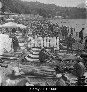 Ein bemerkenswertes Archivfoto aus dem Jahr 1974, das einen unglaublich überfüllten Kiesstrand in Gurzuf, Krim, in der Nähe des internationalen Pioniercamps Artek feststellt. Die Szene ist voll von Hunderten von Urlaubern, die sich unter der Sommersonne auf Holzliegen und Handtüchern sonnen. Im Hintergrund sind die malerischen Hügel des Kurorts und die weiße Promenade-Balustrade deutlich sichtbar. Dieser authentische Vintage-Schnappschuss bewahrt perfekt die geschäftige Urlaubsatmosphäre und den sozialen Lebensstil der sowjetischen Bürger während eines Sommerurlaubs in den 70er Jahren vor Beginn der Moderne. Stockfoto