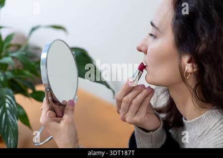 Eine Frau trägt roten Lippenstift auf, während sie einen Spiegel hält, Eine Frau trägt roten Lippenstift auf und hält einen kleinen runden Spiegel vor sich. Stockfoto