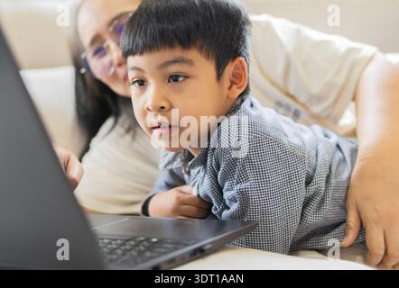 Lächelnde asiatische Mutter und süßer kleiner Sohn liegen auf dem Sofa, während sie den Laptop benutzen und auf den Bildschirm schauen. Stockfoto