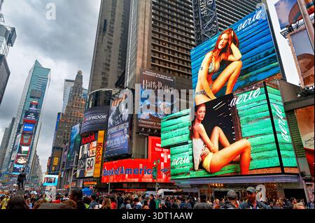 Die Leute laufen und versammeln sich am Times Square. Helle Plakatwände zeigen farbenfrohe Werbung. Die Szene ist mit vielen Besuchern und Einheimischen beschäftigt. Stockfoto