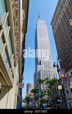 Die Menschen schlendern durch Manhattan, während das Empire State Building unter einem klaren blauen Himmel über ihnen thront. Viele Geschäfte und Gebäude umgeben die Gegend. Stockfoto