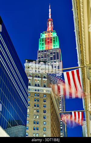 Das Empire State Building steht in New York City hoch und wird in der Abenddämmerung in Farben beleuchtet. Fahnen winken unten gegen den Abendhimmel. Stockfoto