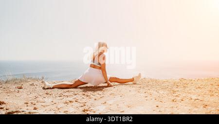 Frau teilt sich die Klippe und führt an einem hellen, sonnigen Sommertag flexible sportliche Bewegungen auf einer felsigen Klippe mit Blick auf das blaue Meer durch Stockfoto
