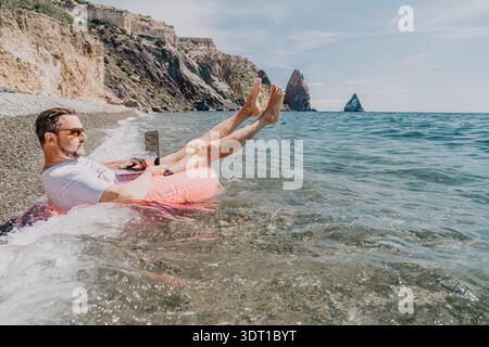 Ein freiberuflicher Laptop Beach man arbeitet an einem aufblasbaren Ring im Ozean, der einen digitalen Nomaden-Lifestyle und ein Remote-Office-Konzept repräsentiert Stockfoto