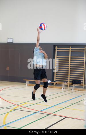 Afroamerikanischer Teenager, der graues T-Shirt trägt, springt und greift in der Turnhalle, um Volleyball zu schlagen Stockfoto