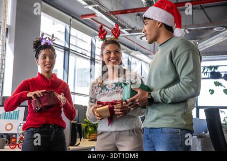 Verschiedene Kollegen tauschen im modernen Büro Geschenke aus, mit Monitor, Weihnachtsmütze und Geweih Stockfoto