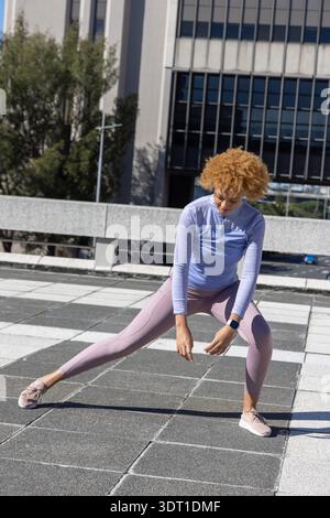 Afroamerikanische Frau, die einen seitlichen Ausfallschritt auf dem gekachelten Dach macht, mit Leggings und Smartwatch Stockfoto