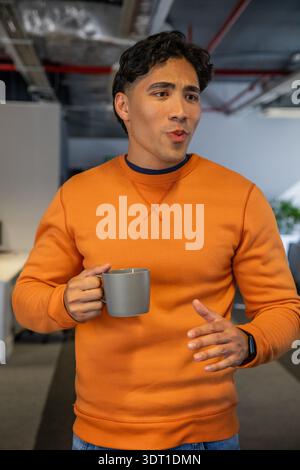 Erwachsener asiatischer Mann im Großraumbüro mit freiliegenden Kanälen, orangefarbenem Rundhalsausschnitt, grauer Becher in der Hand Stockfoto