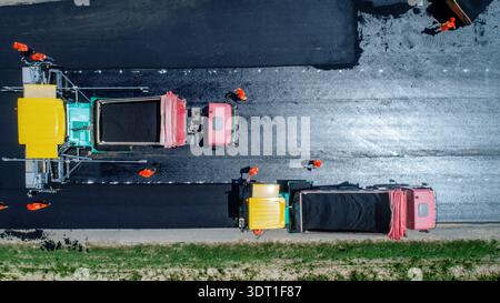 Luftaufnahme von schweren Maschinen, die auf einer neuen Straße dunklen Asphalt verlegen, im Kontrast zu den hellen Maschinen und Arbeitern, Ruma, Vojvodina, Serbien. Stockfoto