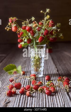 Wilde Erdbeeren in Glasvase auf rustikalem Holztisch, frische Waldbeeren mit grünen Blättern. Natürliches Konzept der Sommerernte, Bio-Obst der Saison Stockfoto