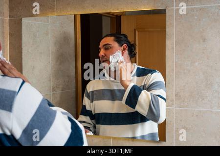 Mann im mittleren Erwachsenenalter, der sich mit der morgendlichen Routine beschäftigt und Rasiercreme auf seine Wange aufträgt, die im Spiegel reflektiert Stockfoto