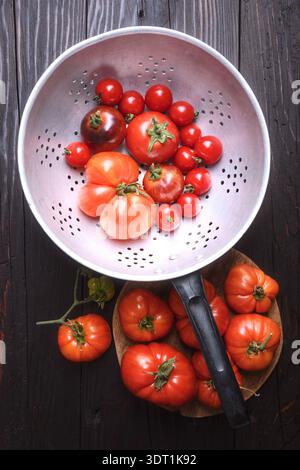 Kirsch- und Erbstück-Tomaten im Sieb mit Messer auf dunklem Holzhintergrund. Frische Bauernprodukte, gesunde Kochzutaten Stockfoto