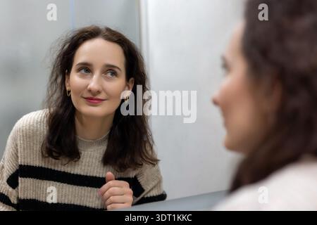 Frau mittleren Alters lächelt bei Spiegelspiegelung im Bad. Nahporträt einer brünetten Frau mittleren Alters, die ihr Spiegelbild im Badezimmer ansieht Stockfoto