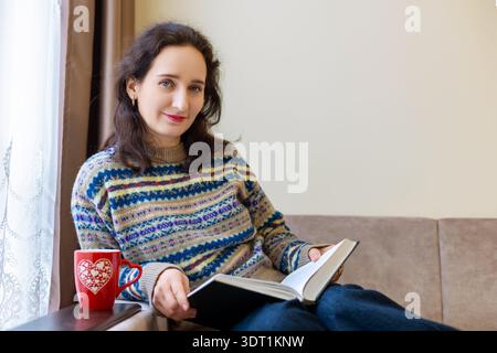 Frau mittleren Alters liest auf Sofa mit Kaffee. Eine brünette Frau mittleren Alters in einem gemusterten Pullover, die ein Buch auf einem Sofa in der Nähe eines Fensters liest, mit einem roten Stockfoto