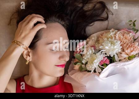 Entspannte Frau, die mit einem Blumenstrauß liegt. Sanfte romantische Nahaufnahme einer brünetten Frau, die mit geschlossenen Augen neben einem Pastellstrauß ruht, gemütlich Stockfoto