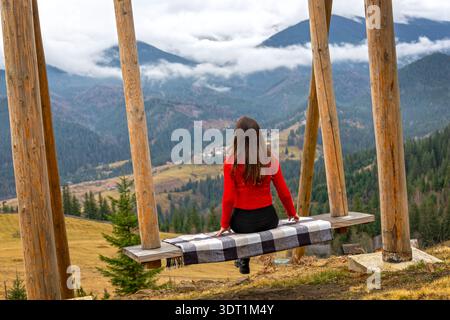 Frau auf Bergschaukel mit Blick auf das nebelige Tal. Frau in rotem Pullover, die auf einer hölzernen Schaukelbank am malerischen Aussichtspunkt mit Blick auf den nebeligen Berg sitzt Stockfoto