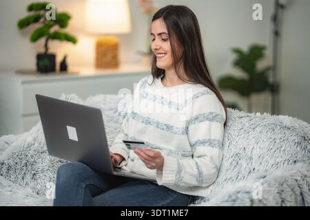 Lächelnde junge Erwachsene Frau beim Online-Shopping mit Laptop und Kreditkarte zu Hause Stockfoto
