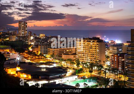 Nächtlicher Blick auf Puerto de la Cruz und beleuchtete Gebäude auf Teneriffa, Kanarischen Inseln, Spanien. Mit den Straßen des Resorts, Palmen und dem Atlantischen Ozean bei Sonnenuntergang. Stockfoto