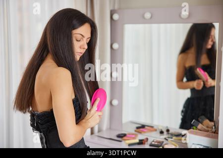 Frau, die vor einem beleuchteten Spiegel steht und ihr langes, dunkles Haar bürstet Stockfoto